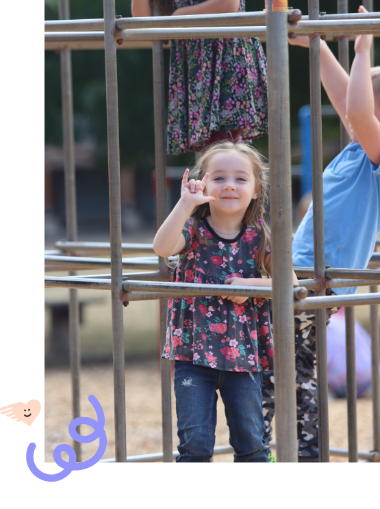 A preschool girl holding up "I love you" fingers and smiling on the jungle gym.