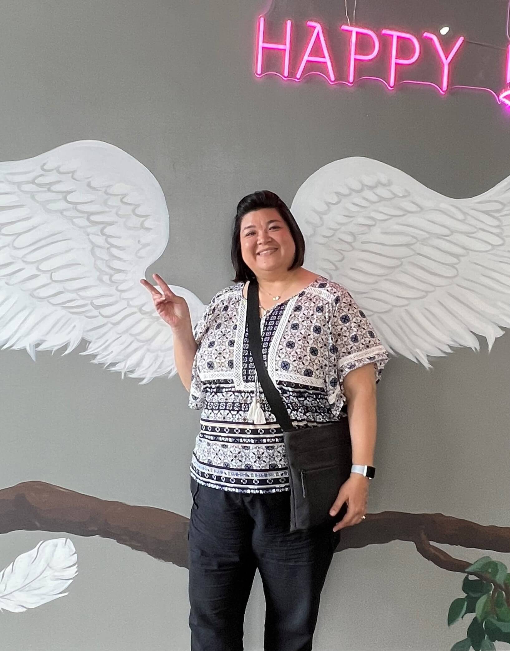 Sheri standing in front of a mural that gives her angel wings. She's smiling and has her fingers in a peace sign.