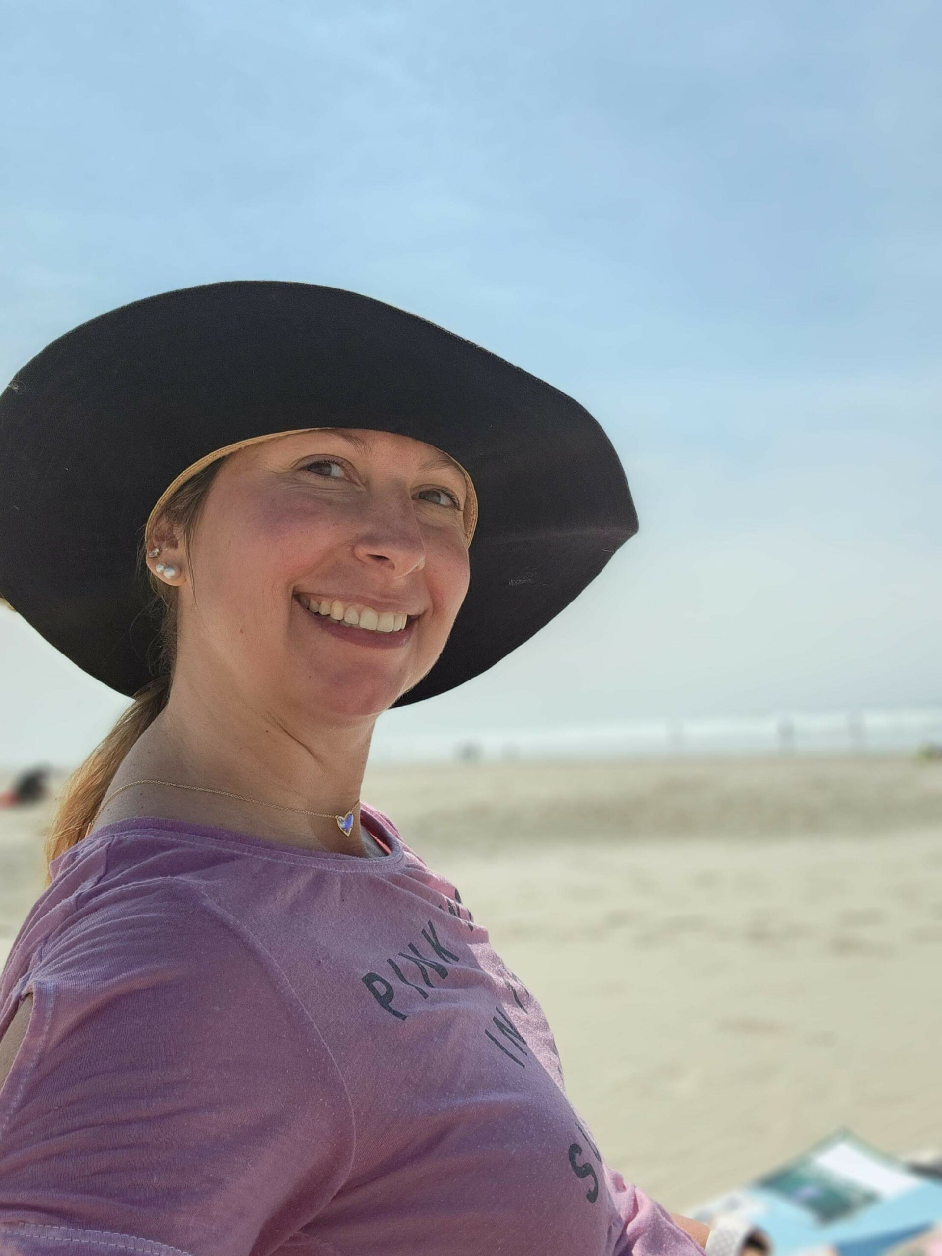 Linda sitting and smiling at the beach. She's wearing a large brimmed black hat.