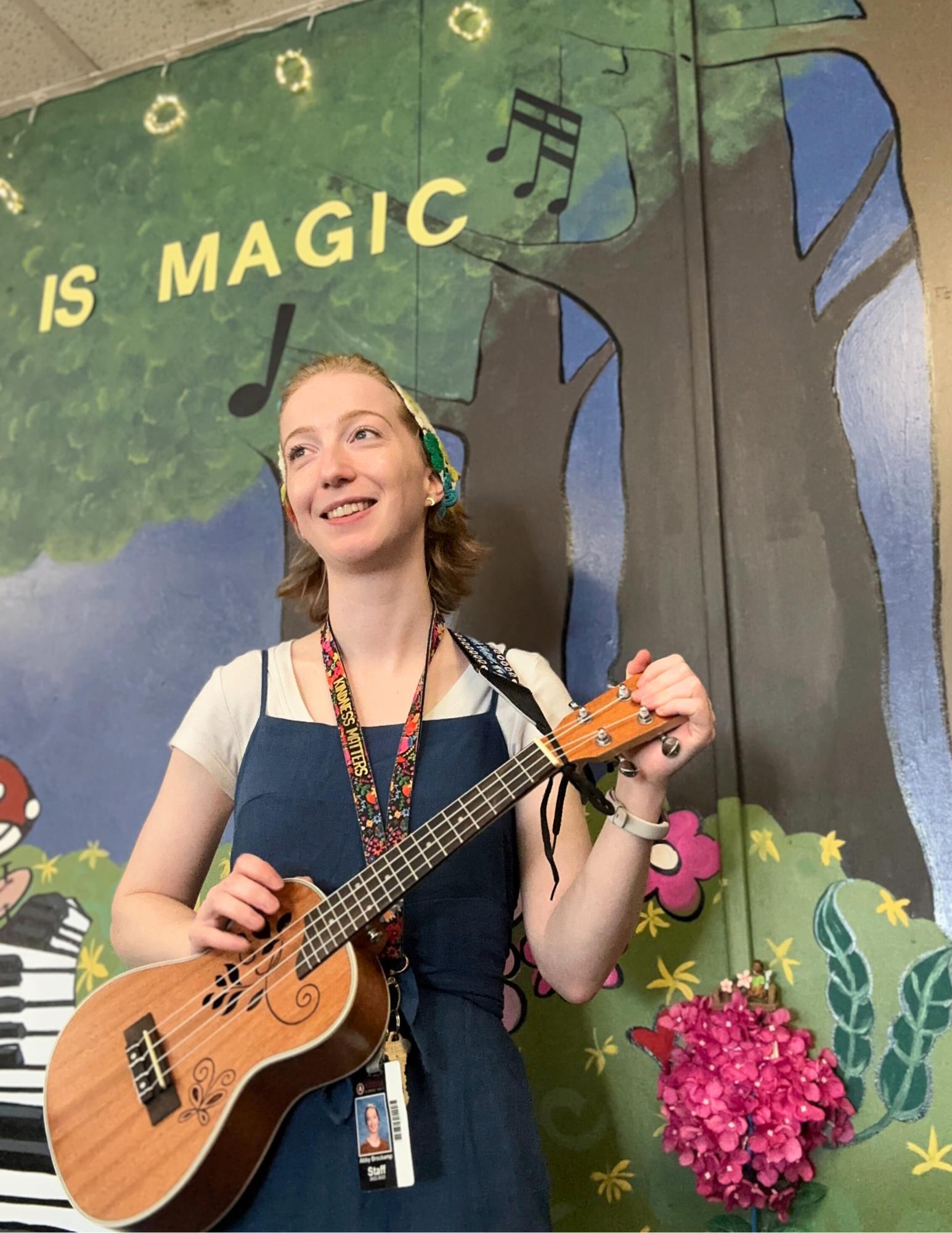 Abby smiling while holding a ukulele in front of her classroom mural