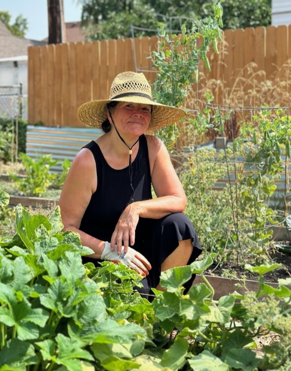 Pam is kneeling in a lush garden, smiling while wearing a large straw hat.